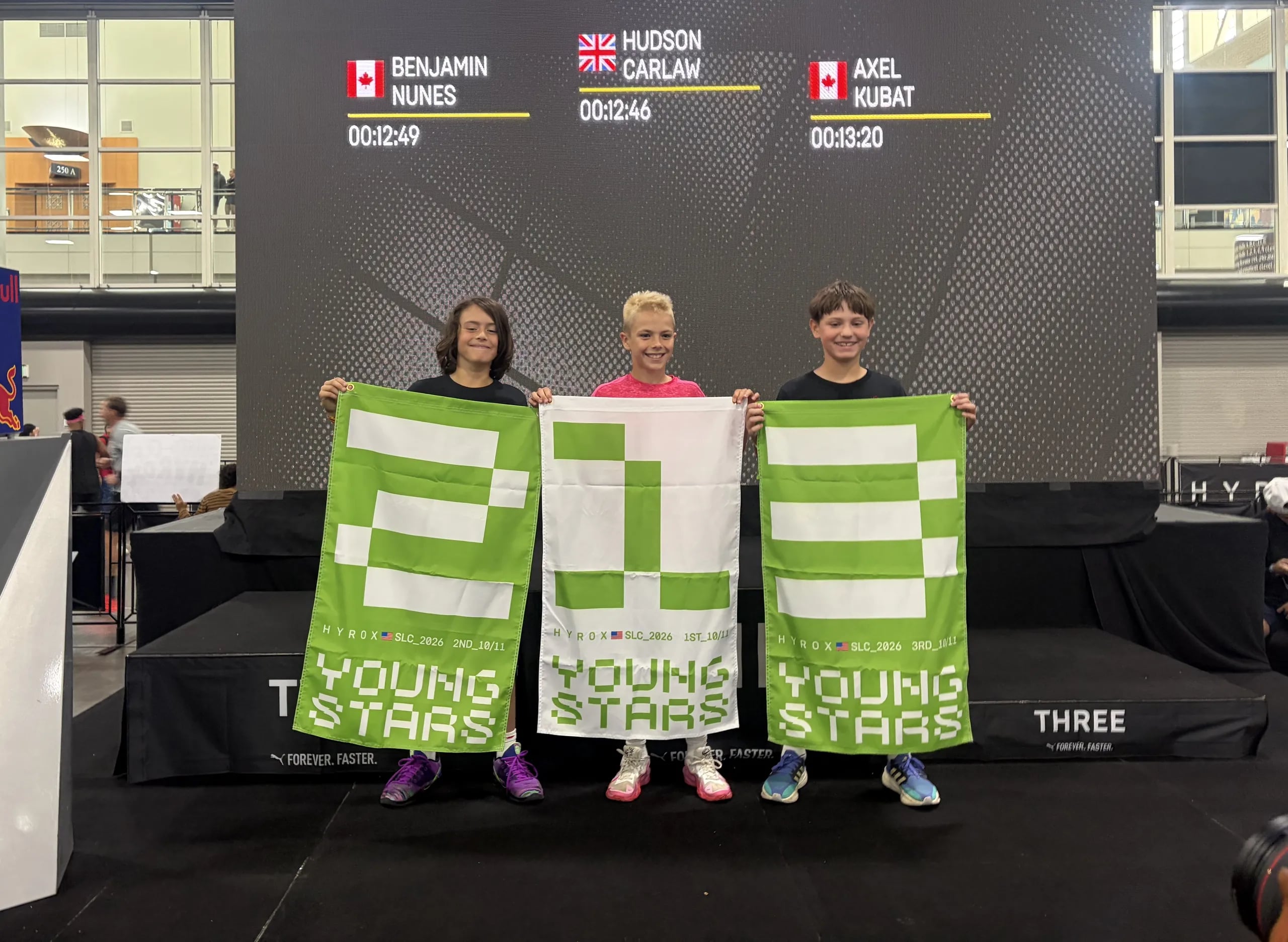 Three young athletes holding 1st, 2nd, and 3rd place HYROX Youngstars flags on the podium at HYROX Salt Lake City 2026.